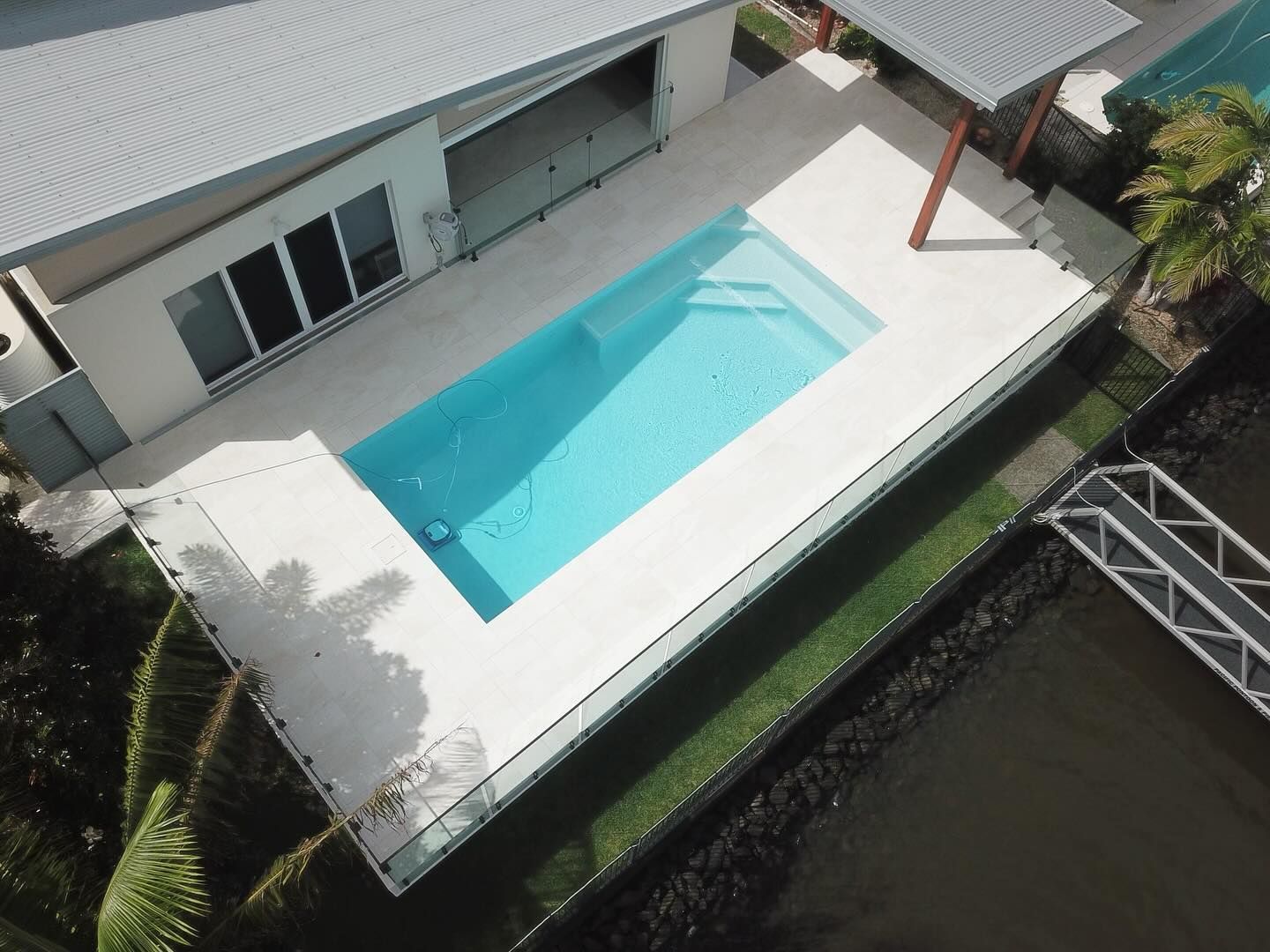 Aerial view of a modern house with a rectangular pool, bright blue water, and a dock on a canal.