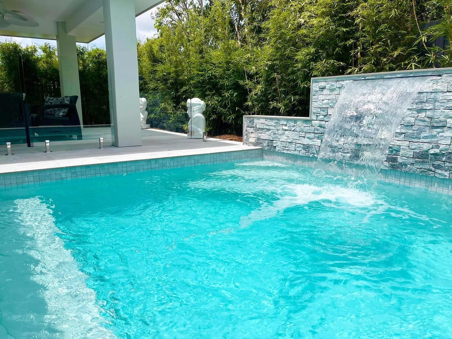 A turquoise pool with a waterfall feature; a covered patio and trees are in the background.