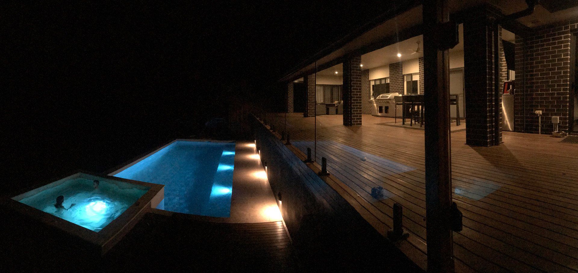 A nighttime view of a pool and jacuzzi lit with blue lights beside a wooden deck and modern house.
