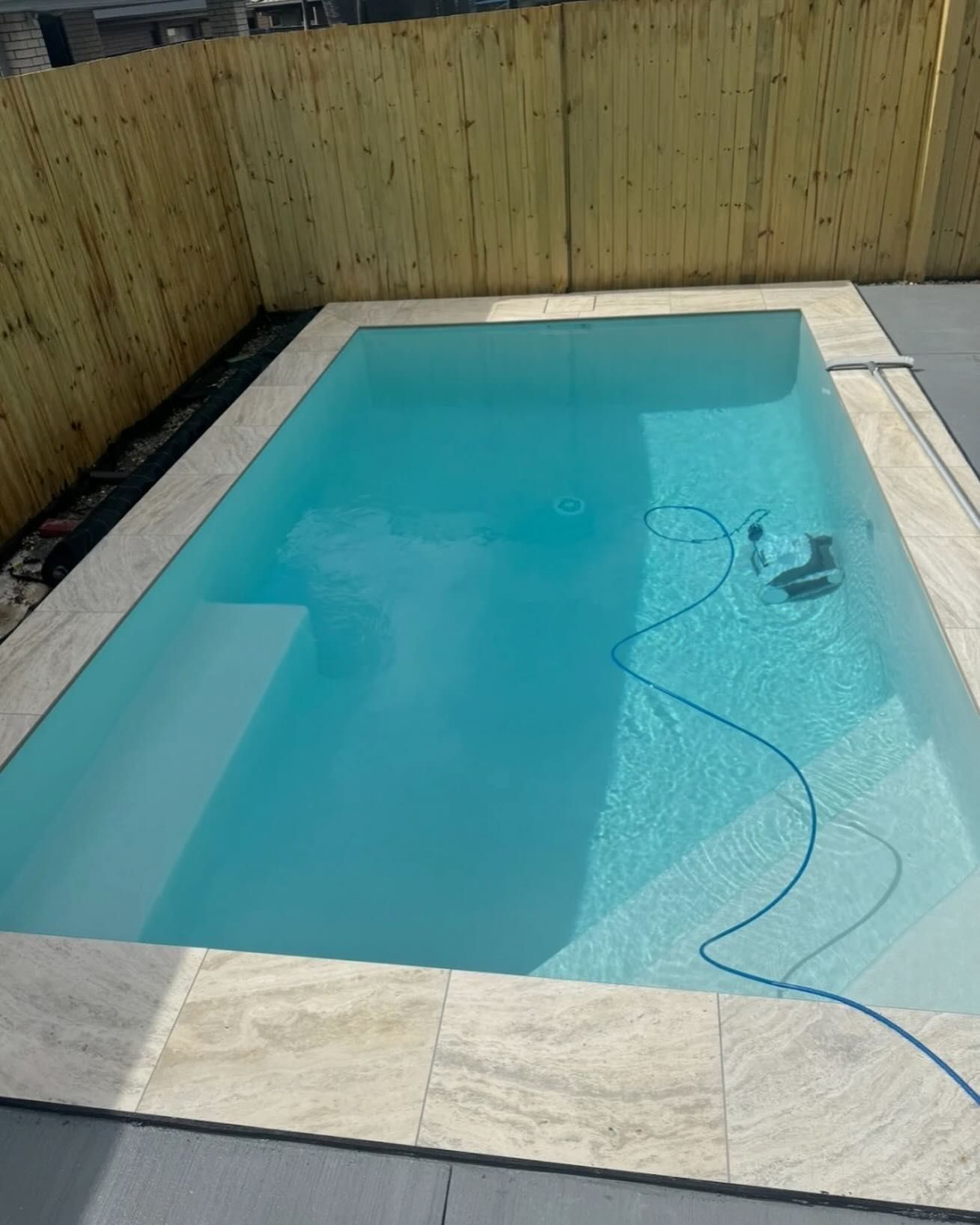 Rectangular swimming pool with light blue water, bordered by beige tiles and a wooden fence.