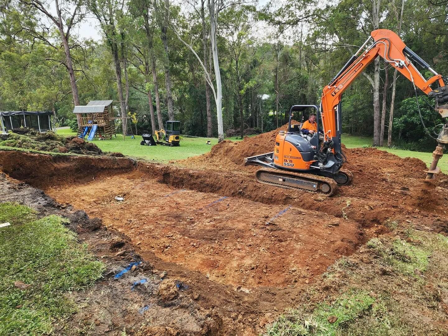 An excavator digging a rectangular hole in a grassy area, another small machine in the background, trees.