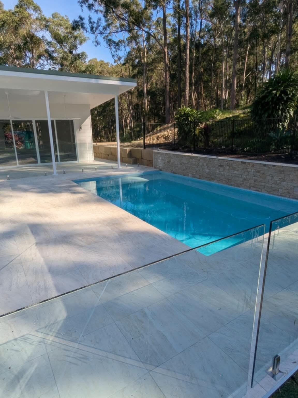 Swimming pool with glass fence and surrounding light-colored stone patio, set against a backdrop of trees.
