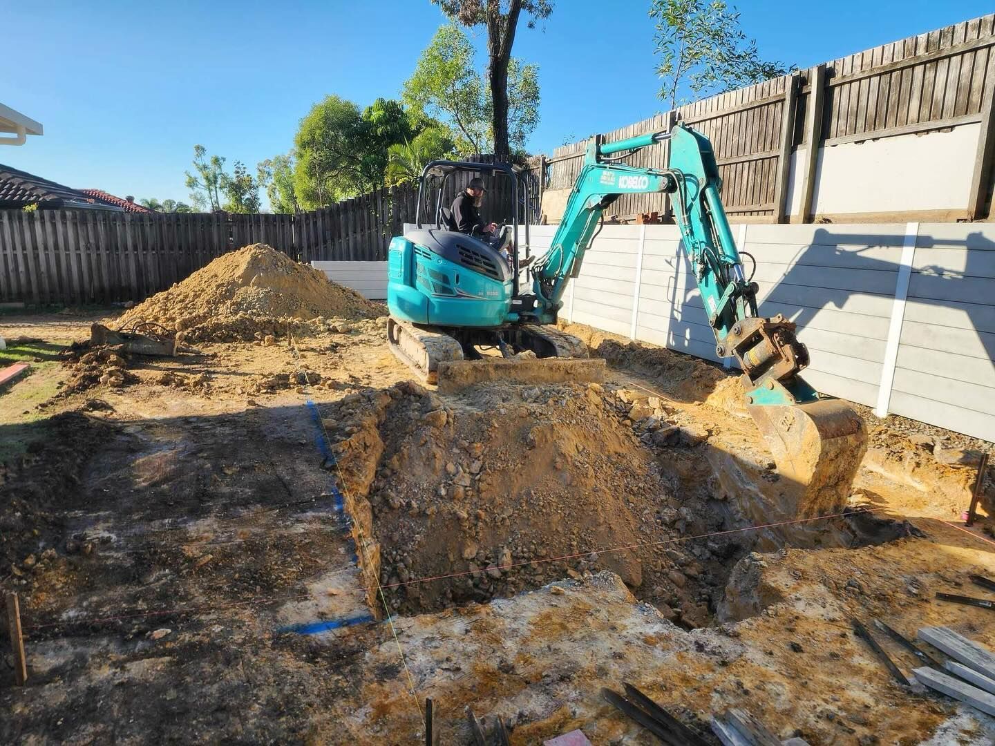 A small teal excavator digging dirt in a backyard with a wooden fence.