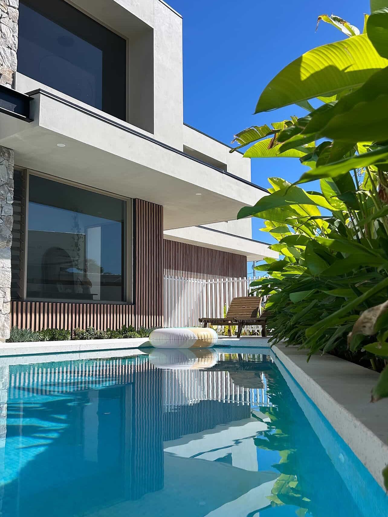 Modern villa with a long pool reflecting the building and the blue sky; lush greenery on the right.