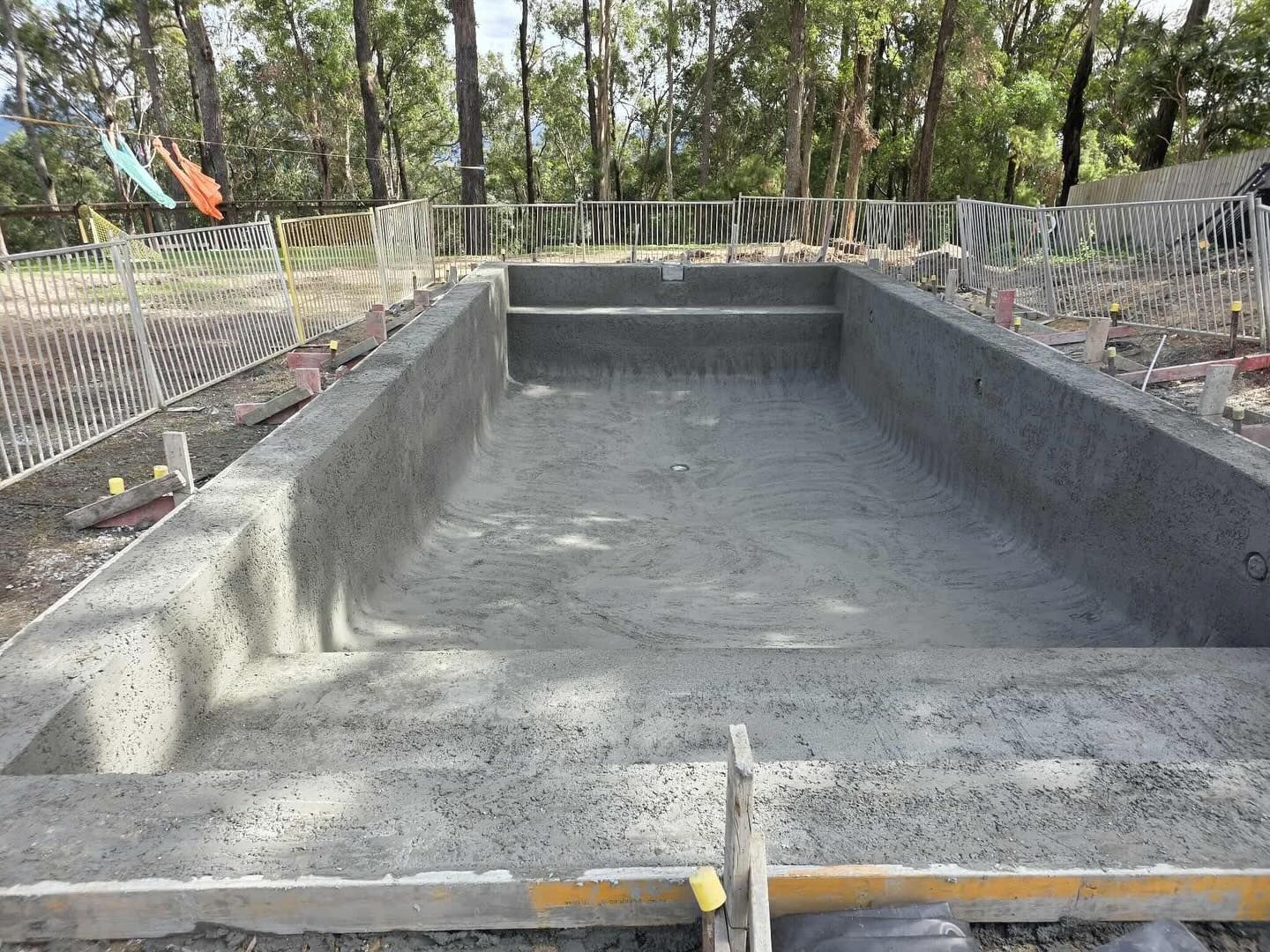 Concrete swimming pool under construction outdoors with steps and surrounding fencing.