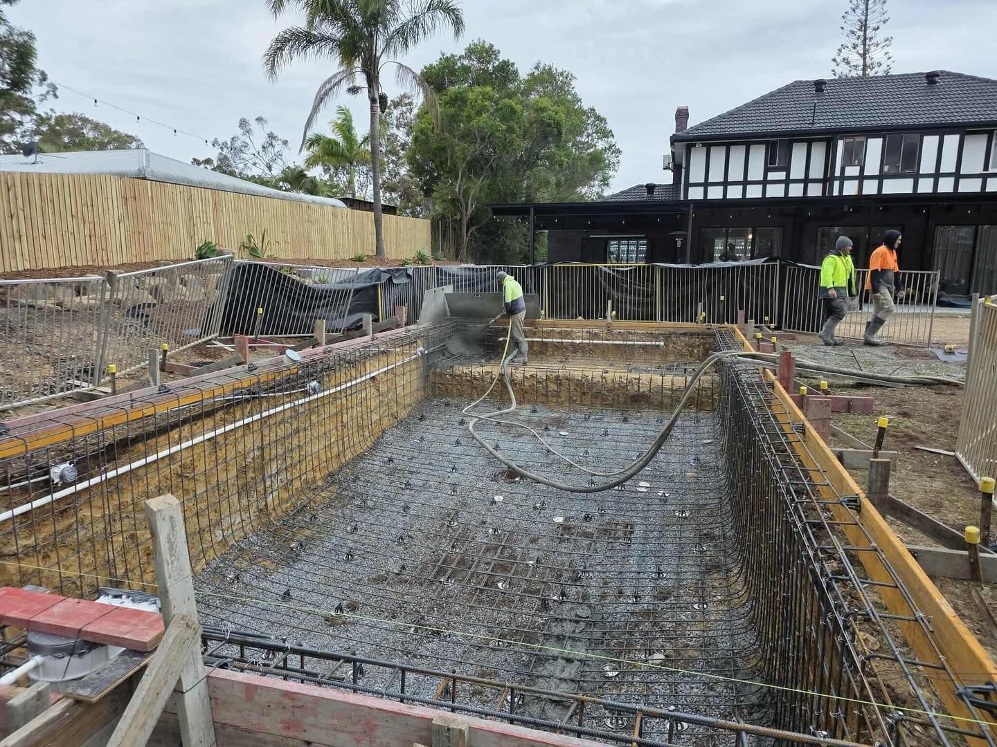 Construction site: workers pouring concrete into a swimming pool form, surrounded by wooden frames and rebar.