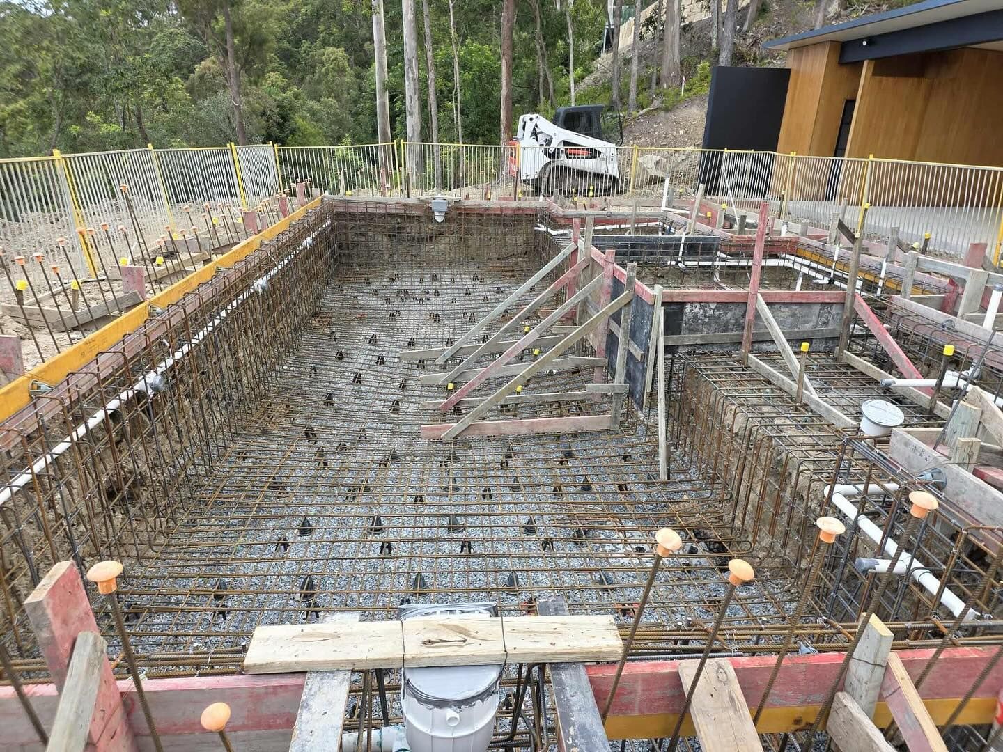 Pool construction: metal framework in excavated rectangular hole, surrounded by wooden forms, outside.