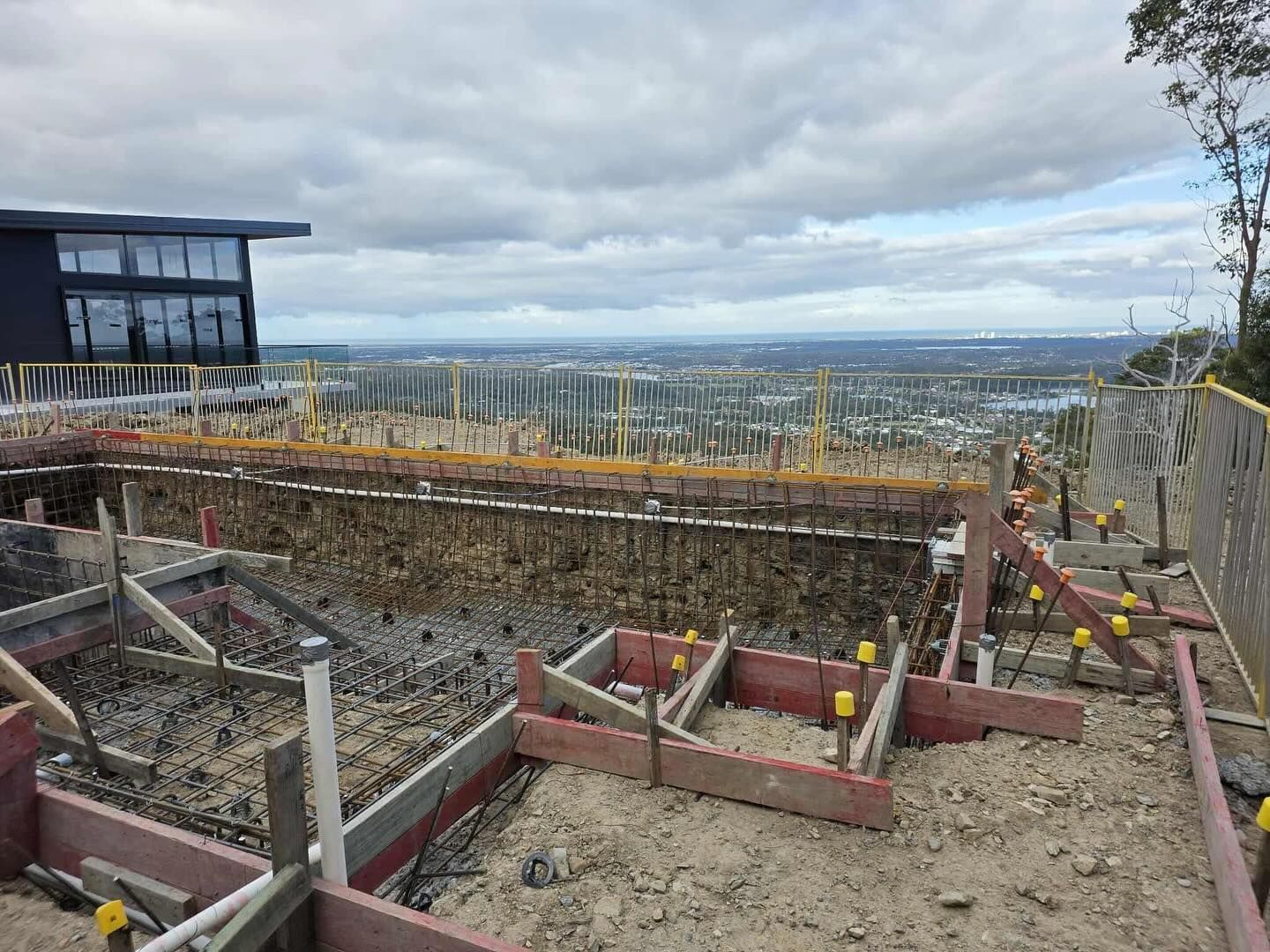 Construction site with concrete forms, rebar, and city view under cloudy sky.