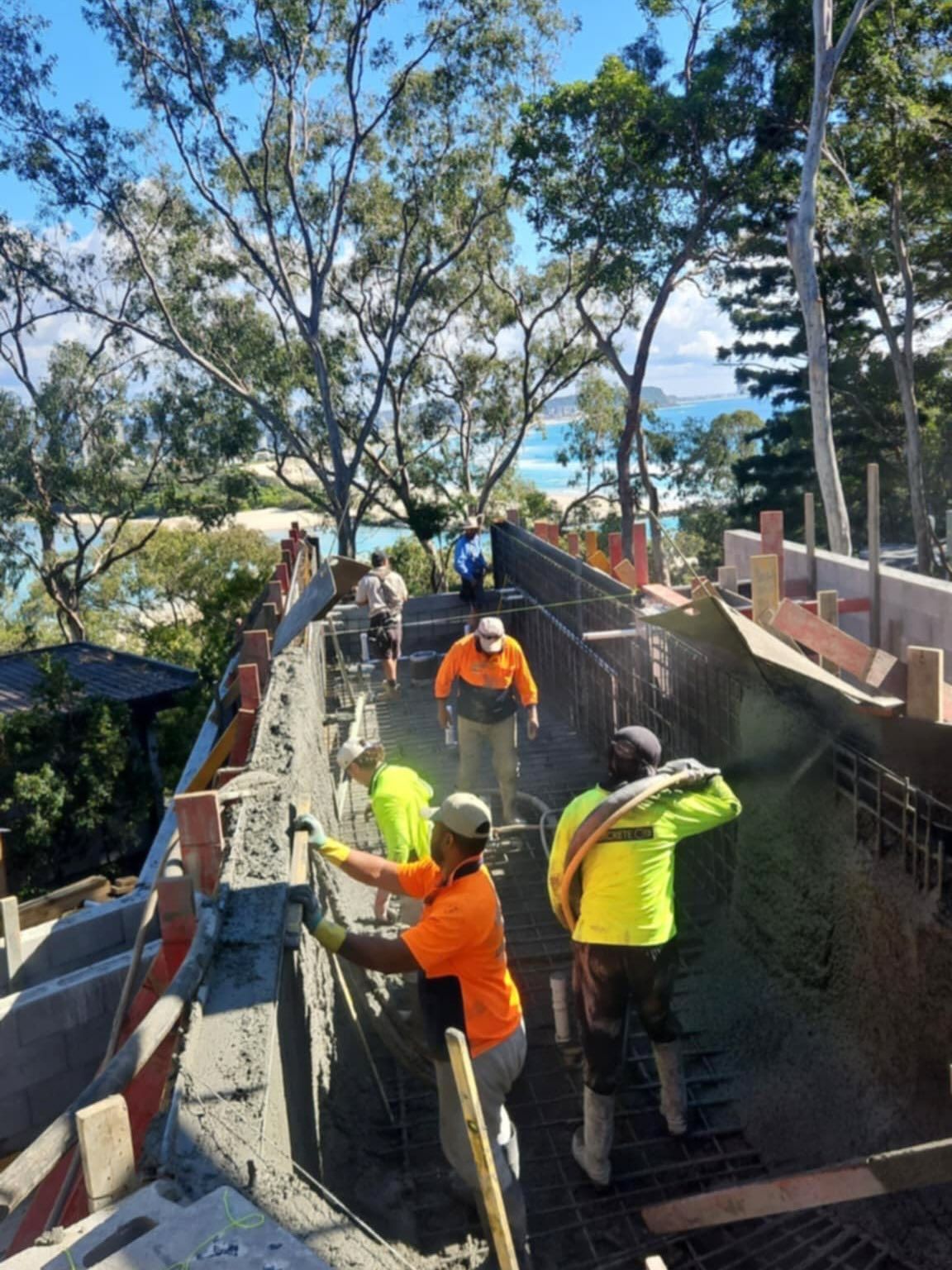 Construction workers pouring concrete on a walkway with ocean view, wearing safety vests and gloves.
