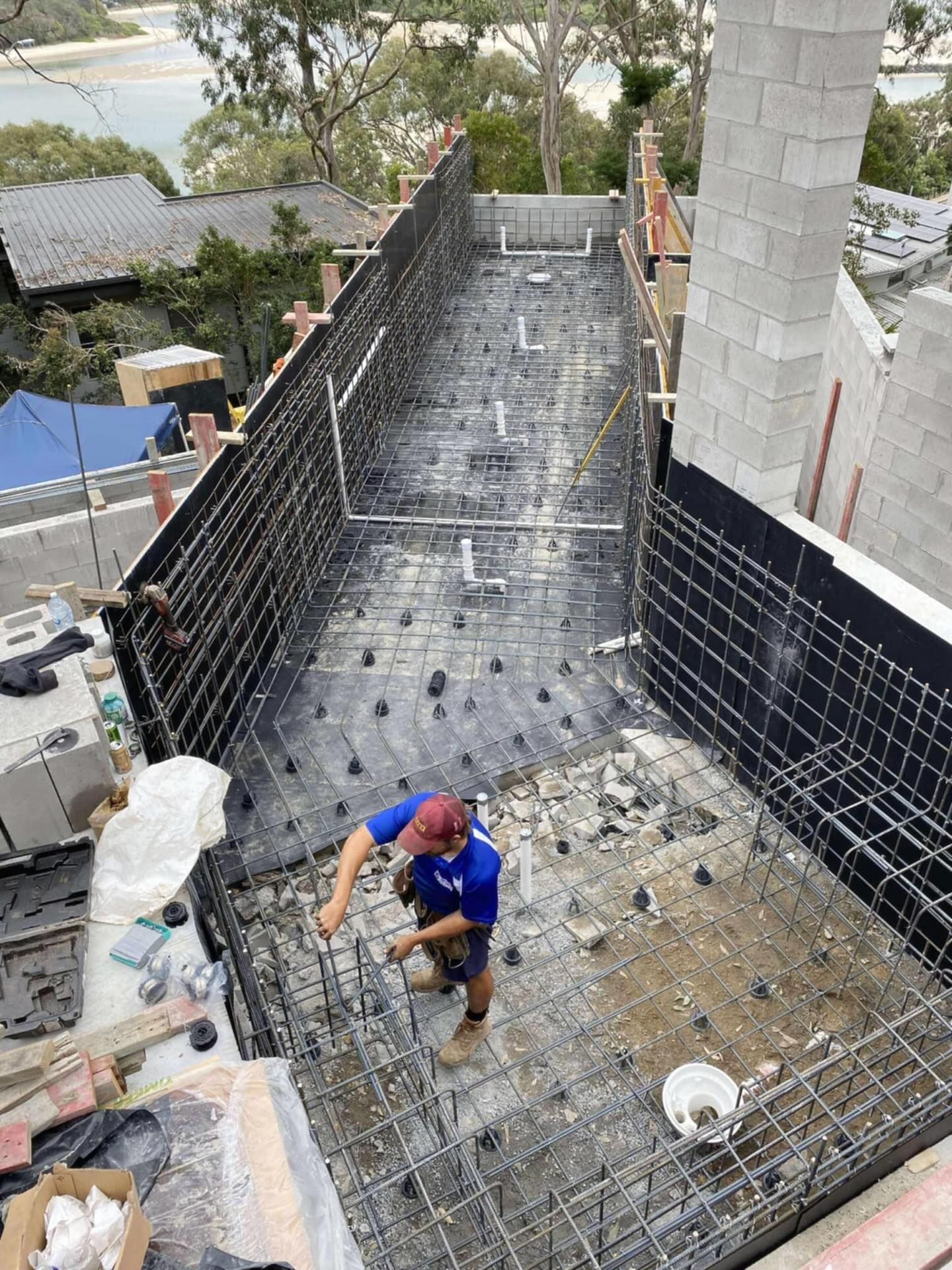 Construction worker tying rebar in a pool being built, black mesh on walls, above a body of water.