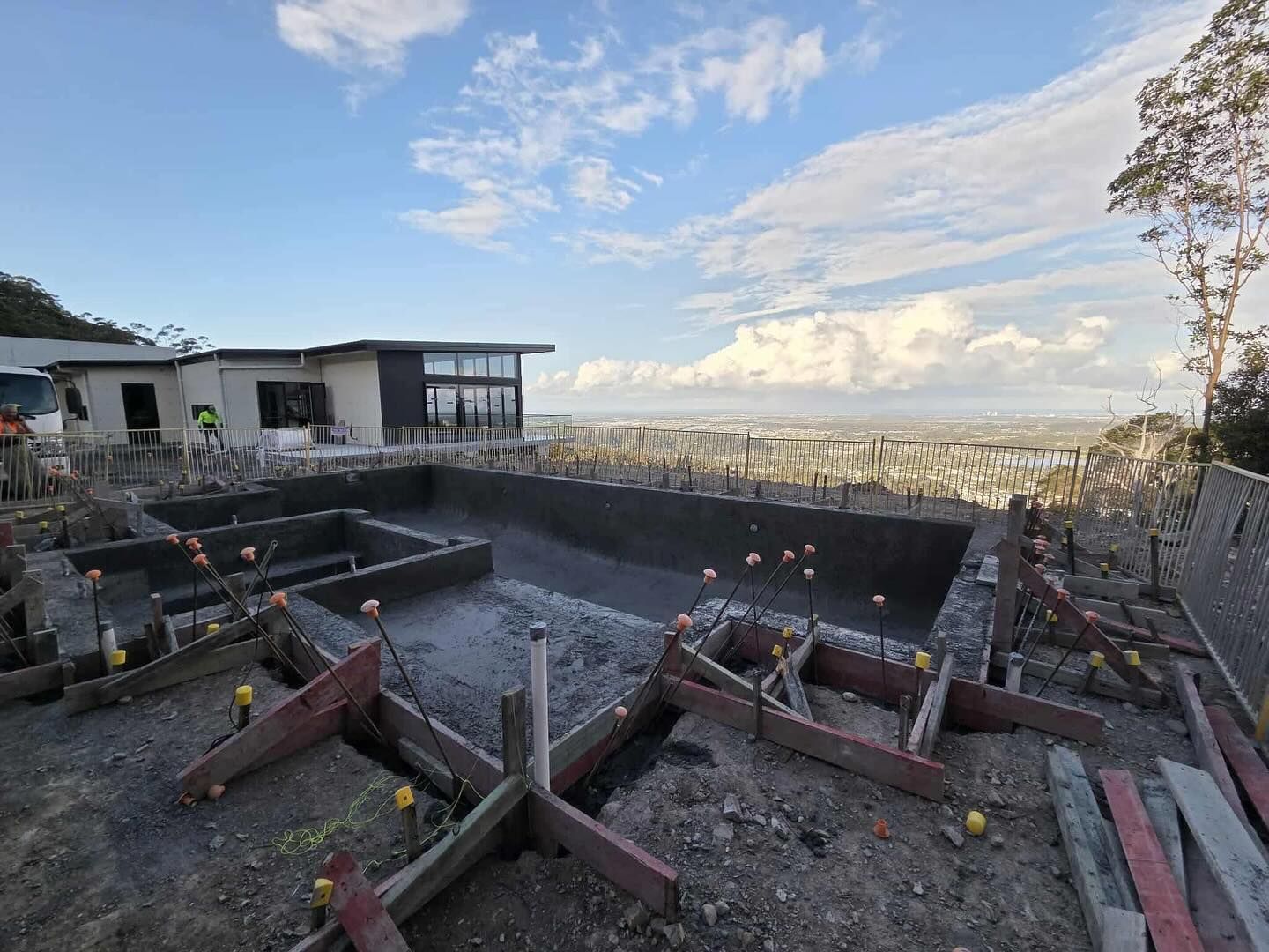Construction site with a pool foundation, house in background, cloudy sky.