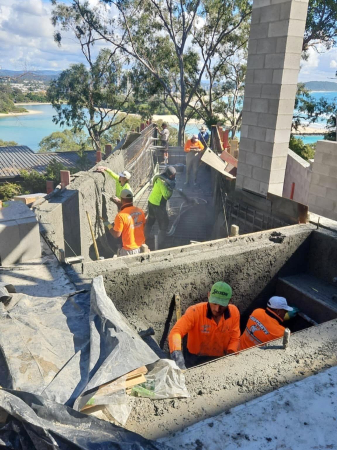 Construction workers pouring concrete into a large outdoor structure with a scenic ocean view.