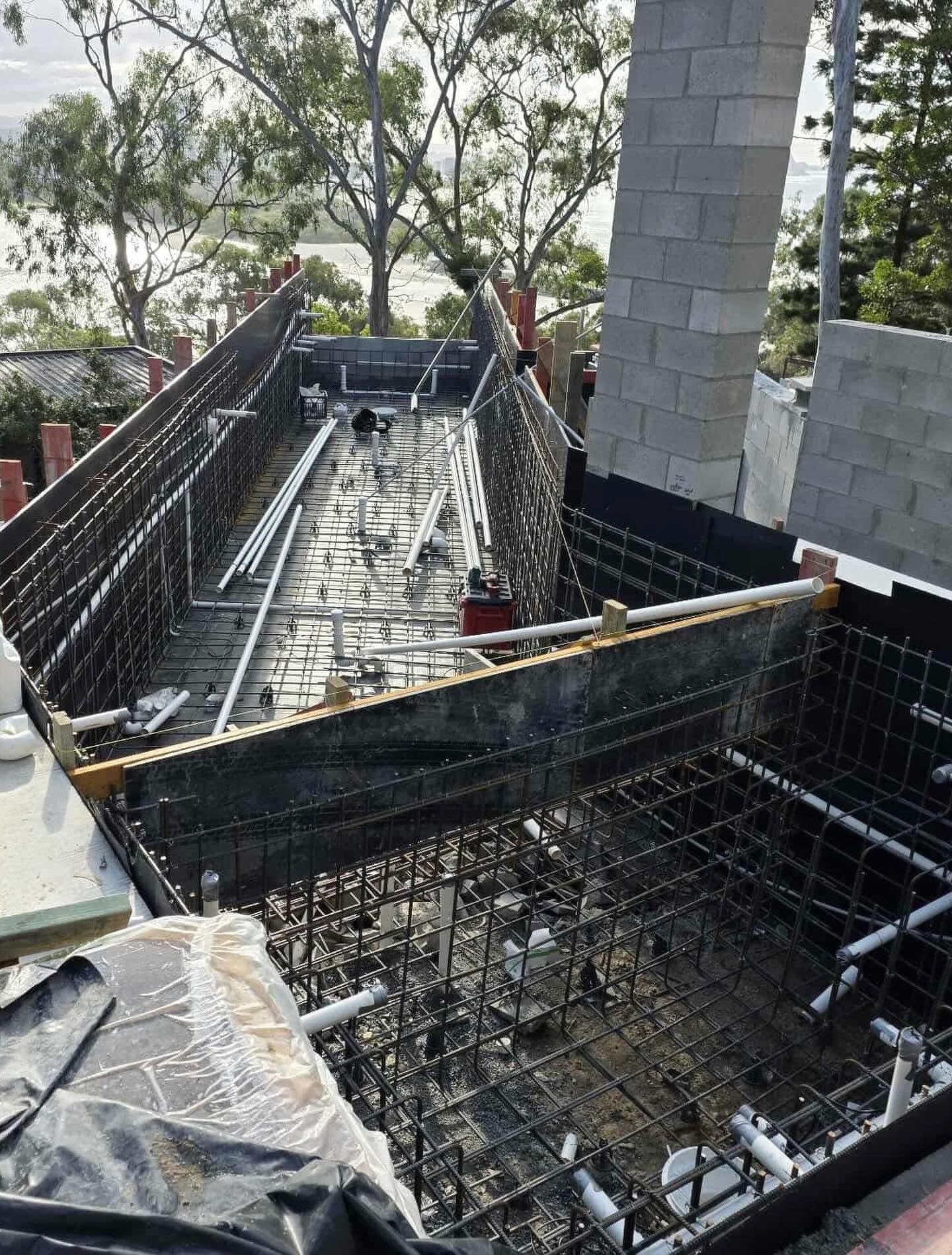 Construction of a swimming pool, with exposed rebar, pipes, and forms; brick wall visible.