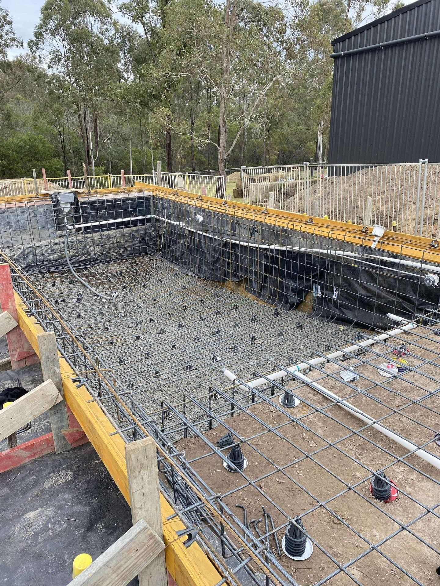 Pool construction site with rebar, gravel, and black lining, surrounded by wooden forms and a dark building.