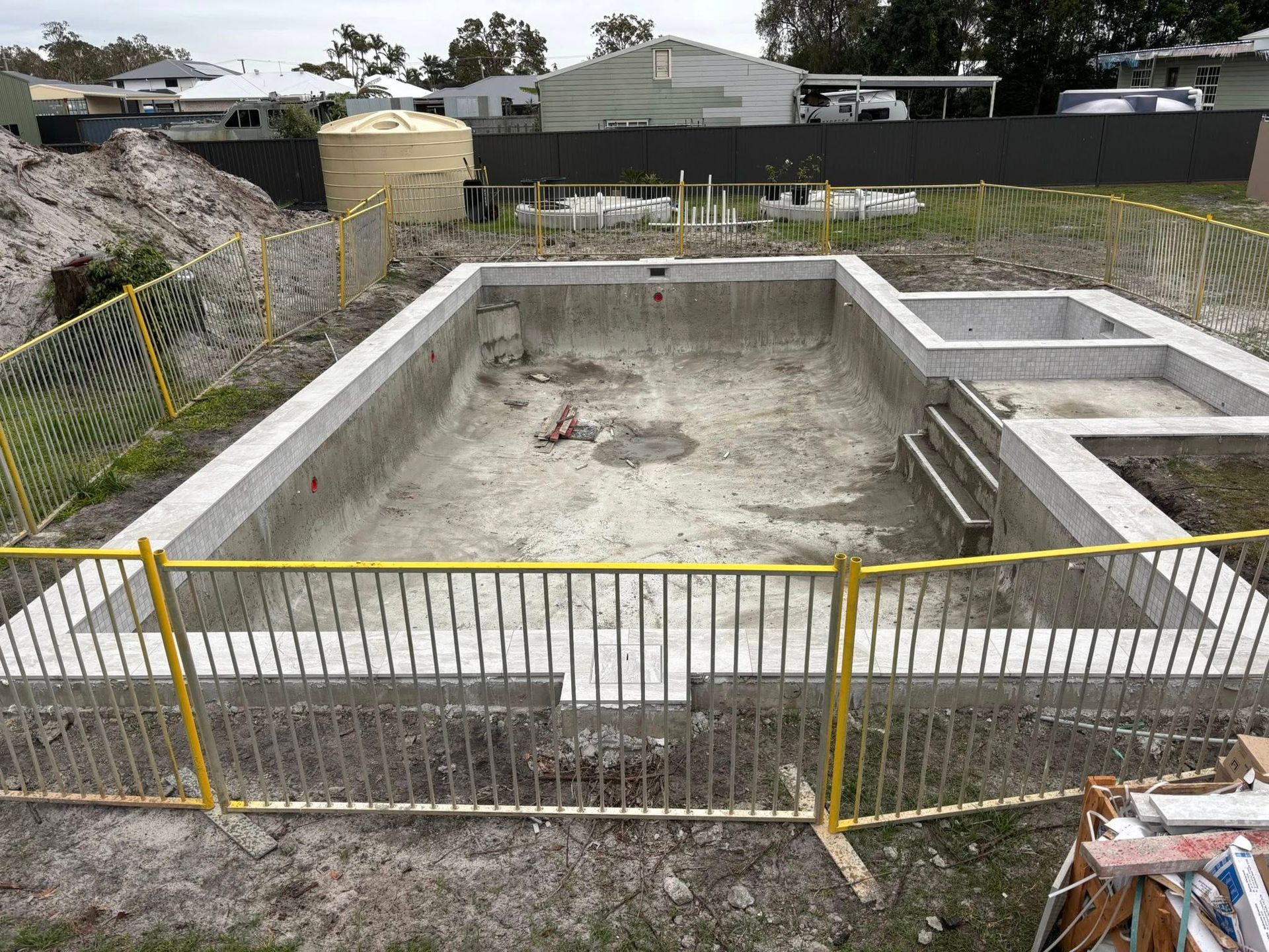 Construction site with a concrete swimming pool under construction, surrounded by a yellow safety fence.