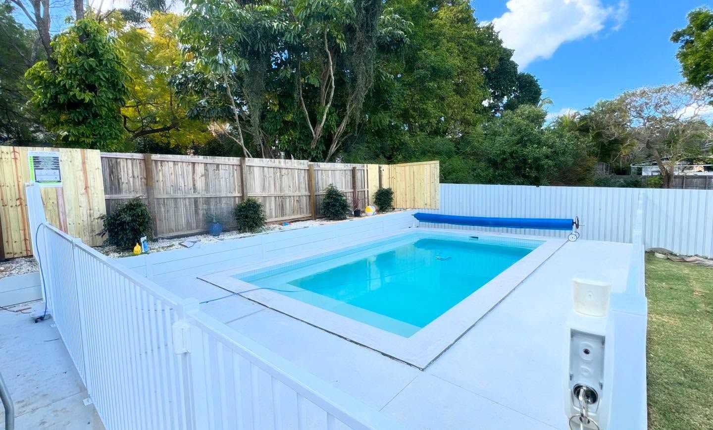 A rectangular pool with blue water in a fenced-in backyard with white decking and a blue pool cover.