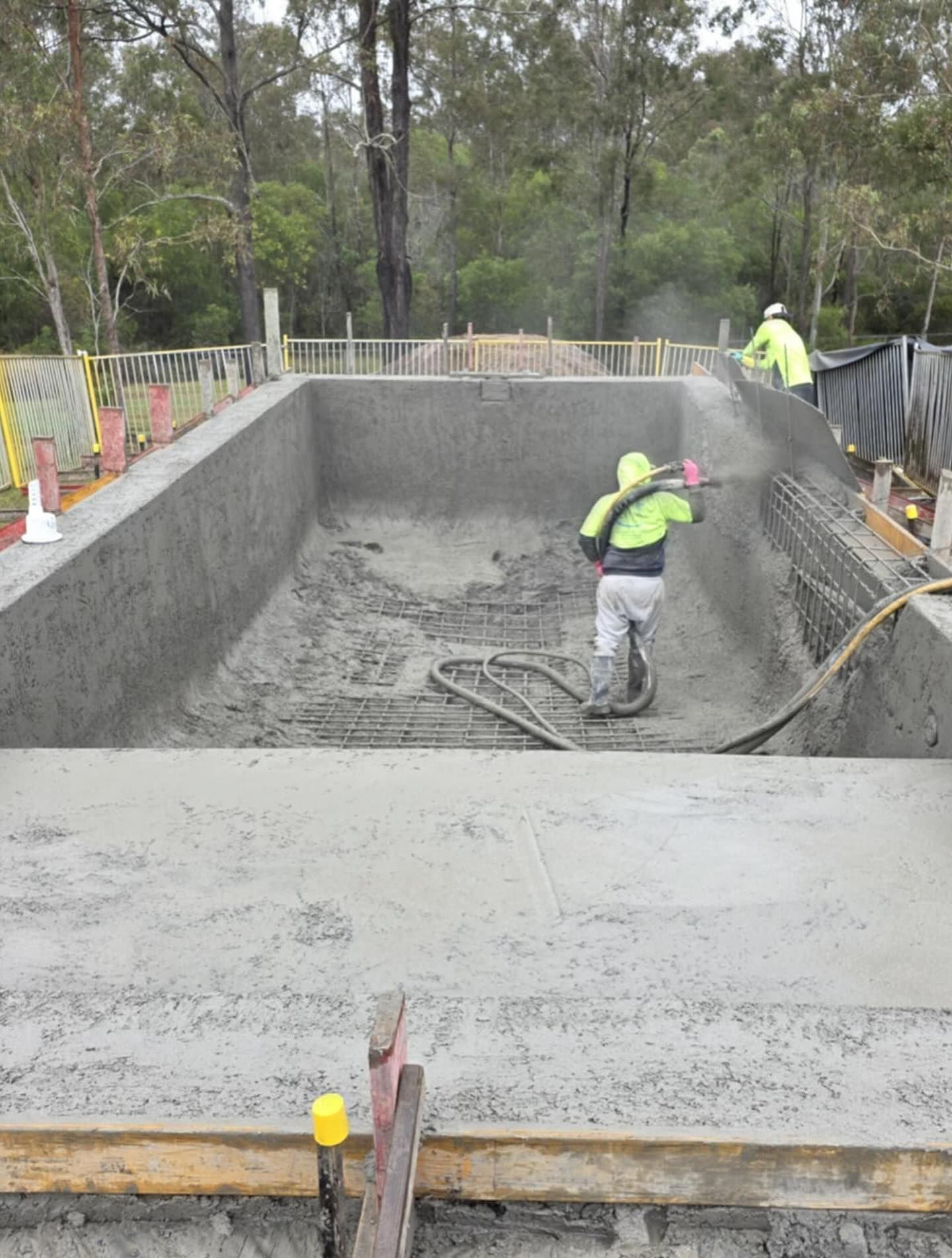 Workers spraying concrete inside a pool under construction; setting is outdoors.