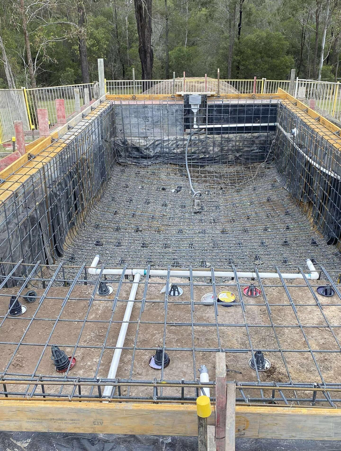 Construction of a rectangular swimming pool. Metal rebar grid with pipes. Wooden forms and surrounding trees.