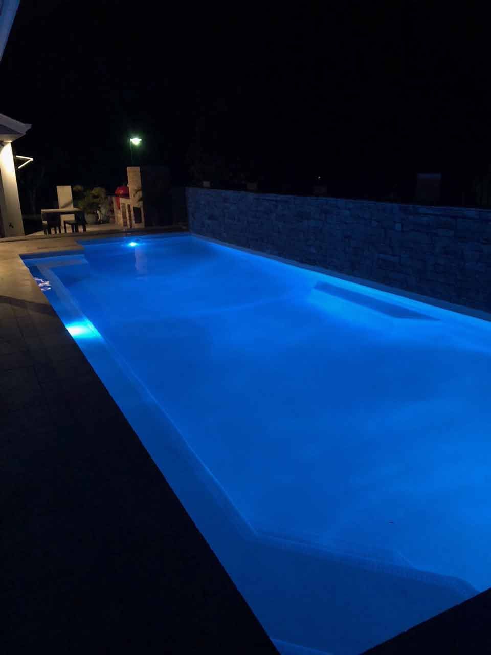 Nighttime view of a blue-lit rectangular swimming pool with a dark sky overhead and patio furniture in the background.