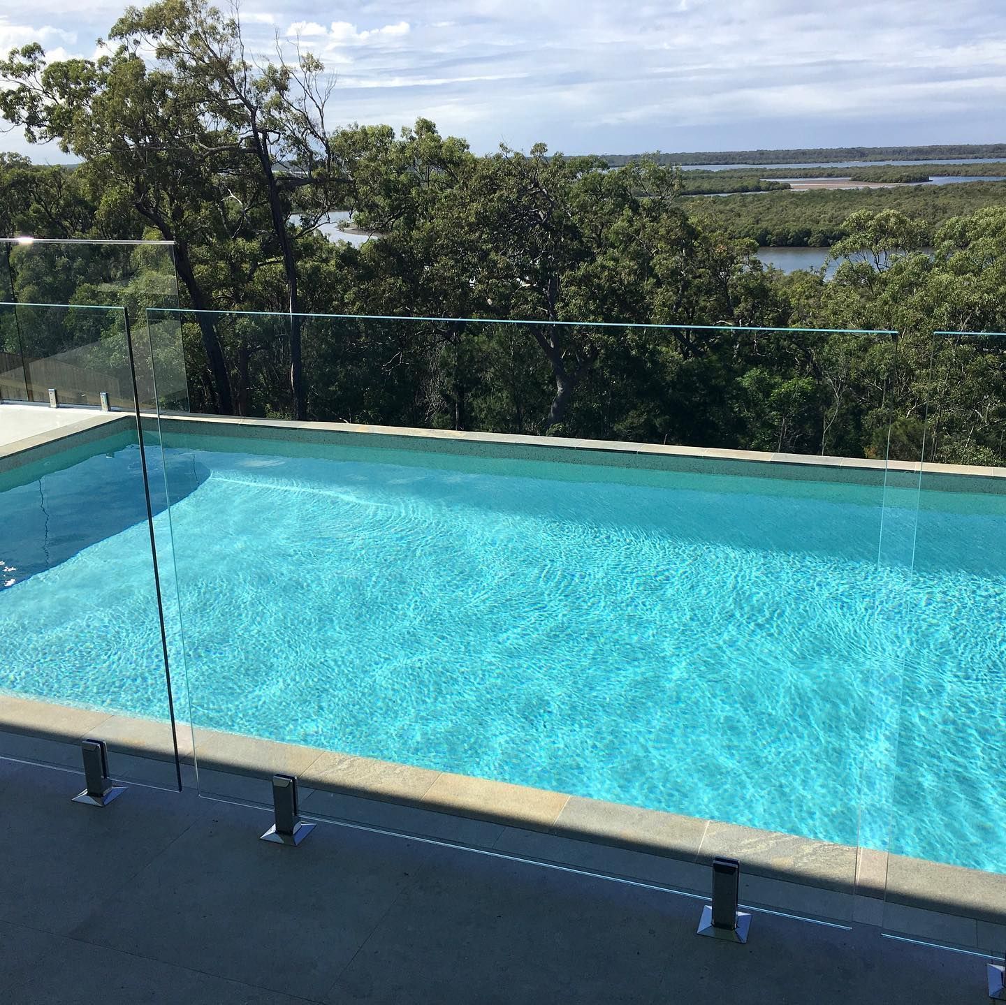 Pool with glass railing overlooking a tree-lined river under a partly cloudy sky.