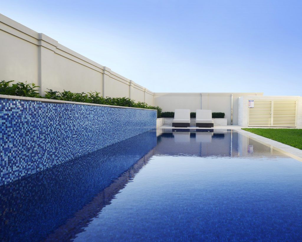Blue-tiled pool reflects the clear sky. A low wall with plants runs alongside the pool.