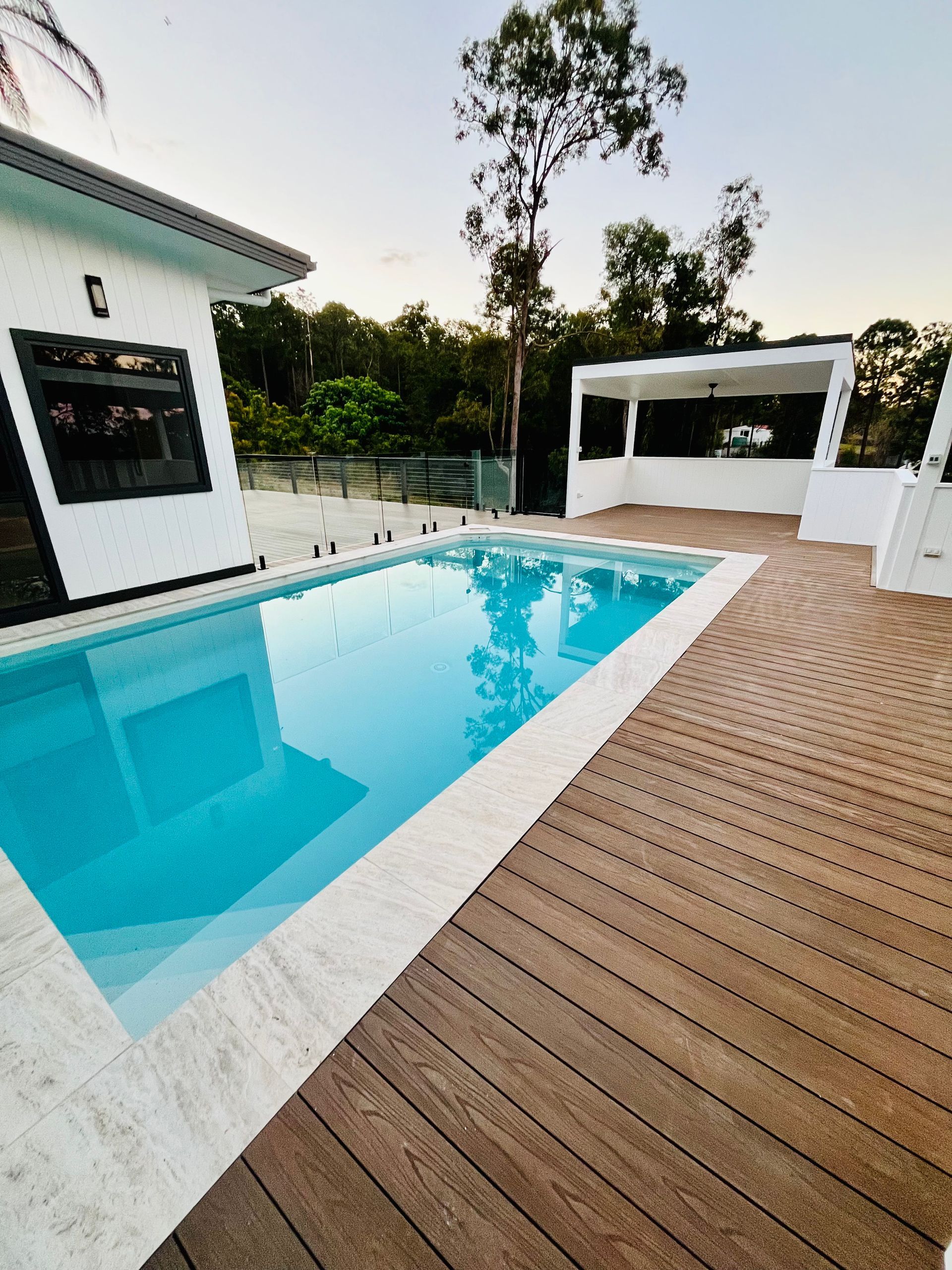 A modern pool with turquoise water and a wooden deck next to a white house and gazebo, set in a wooded area.