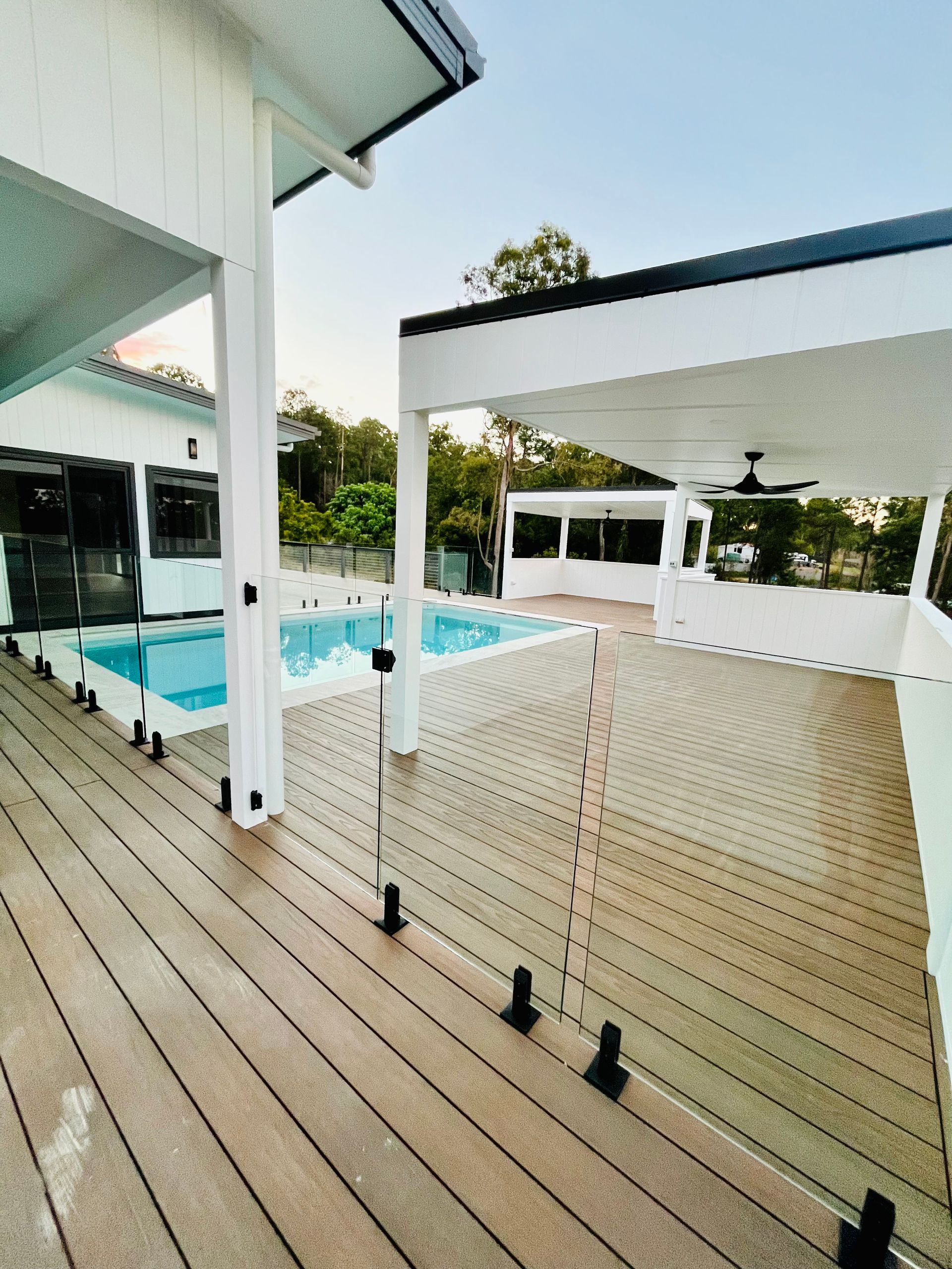 Deck with glass railing overlooks a pool and covered patio; white house with black accents.