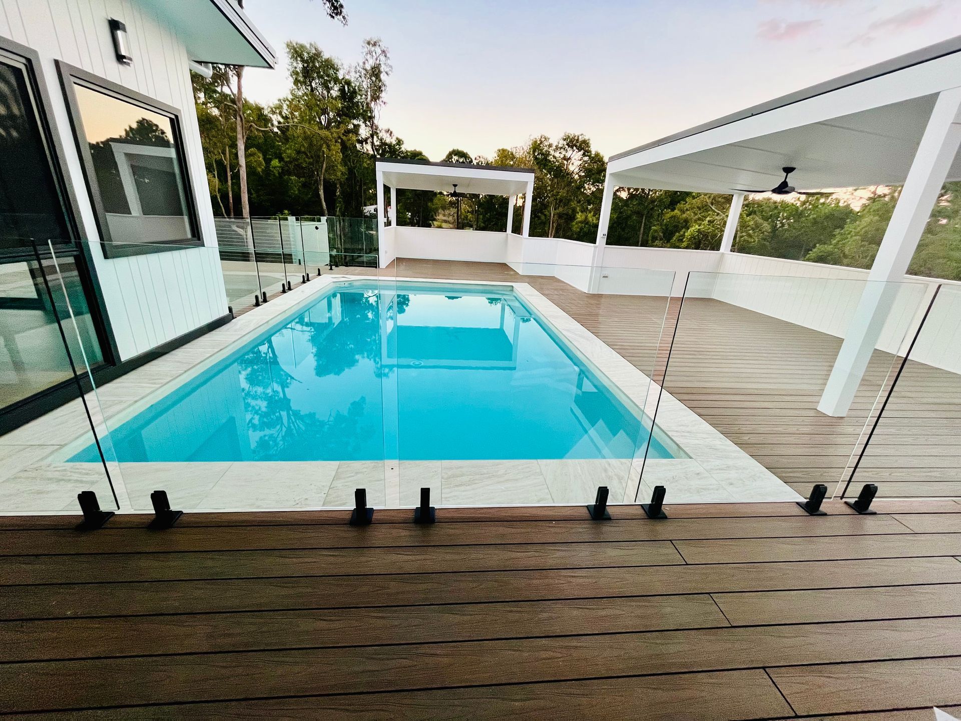 Rectangular pool with clear water, surrounded by a wooden deck and glass fencing, under a white pergola.