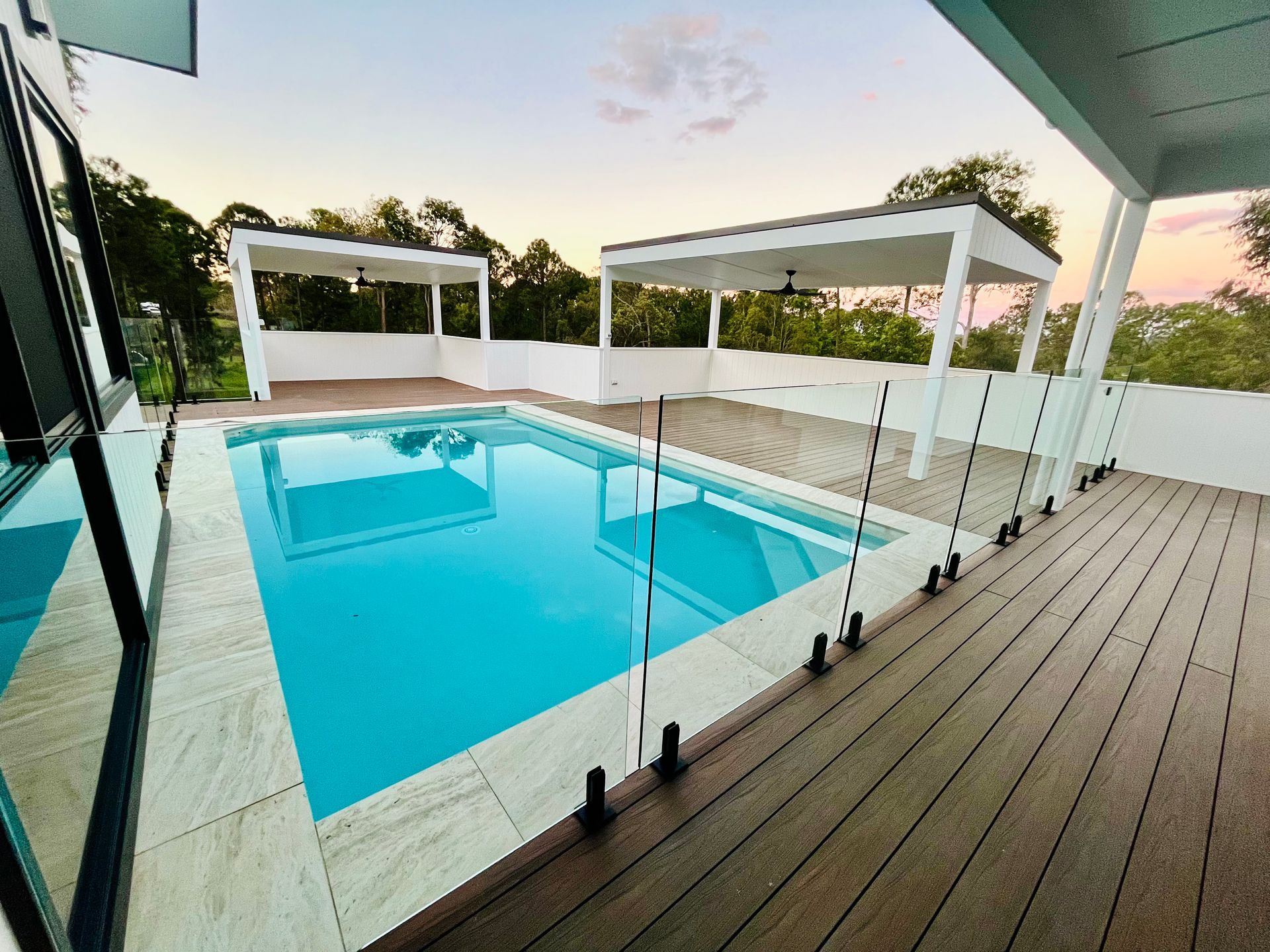 Pool with turquoise water surrounded by a deck and two white pergolas, set against a sunset sky.