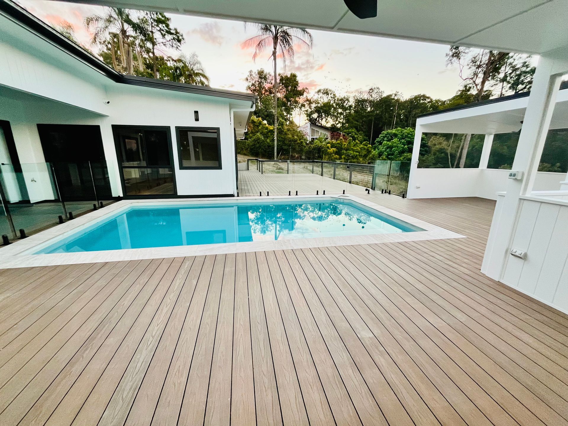 Wooden deck surrounds a blue rectangular pool next to a white house and a small covered area.