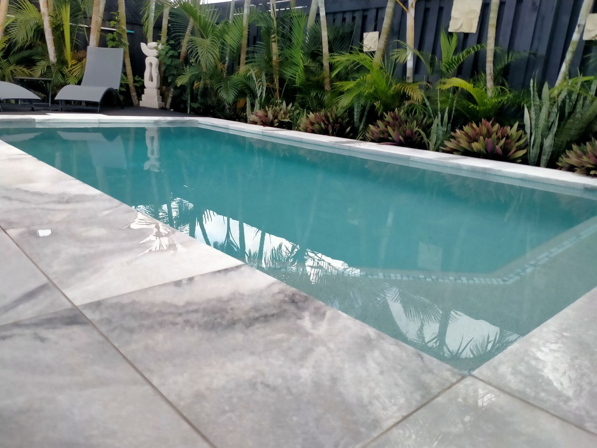 Rectangular pool with gray tile surround, surrounded by lush greenery and a dark fence.