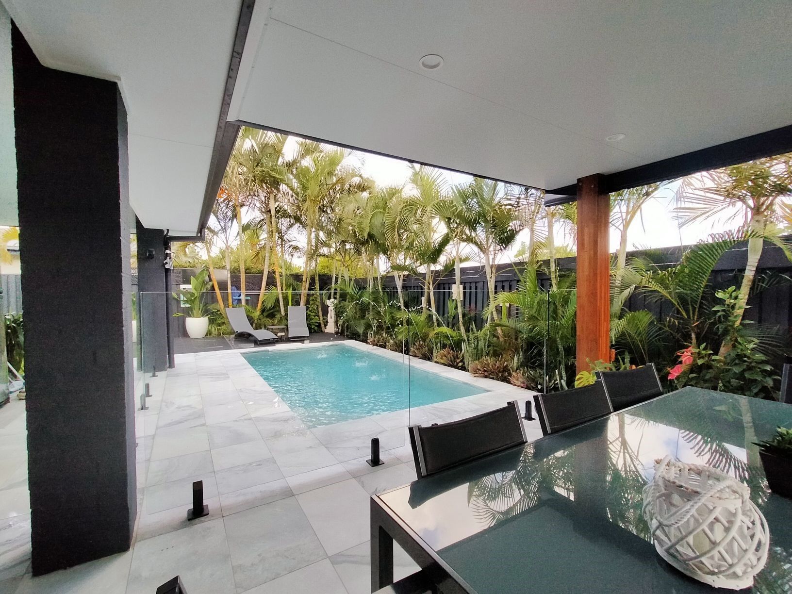 Poolside patio with pool, dining table, and lounge chairs. Lush tropical greenery surrounds the pool.
