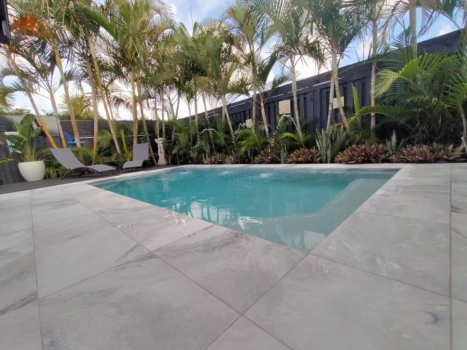 A swimming pool surrounded by light stone tiles, palm trees, and black fences under a blue sky.