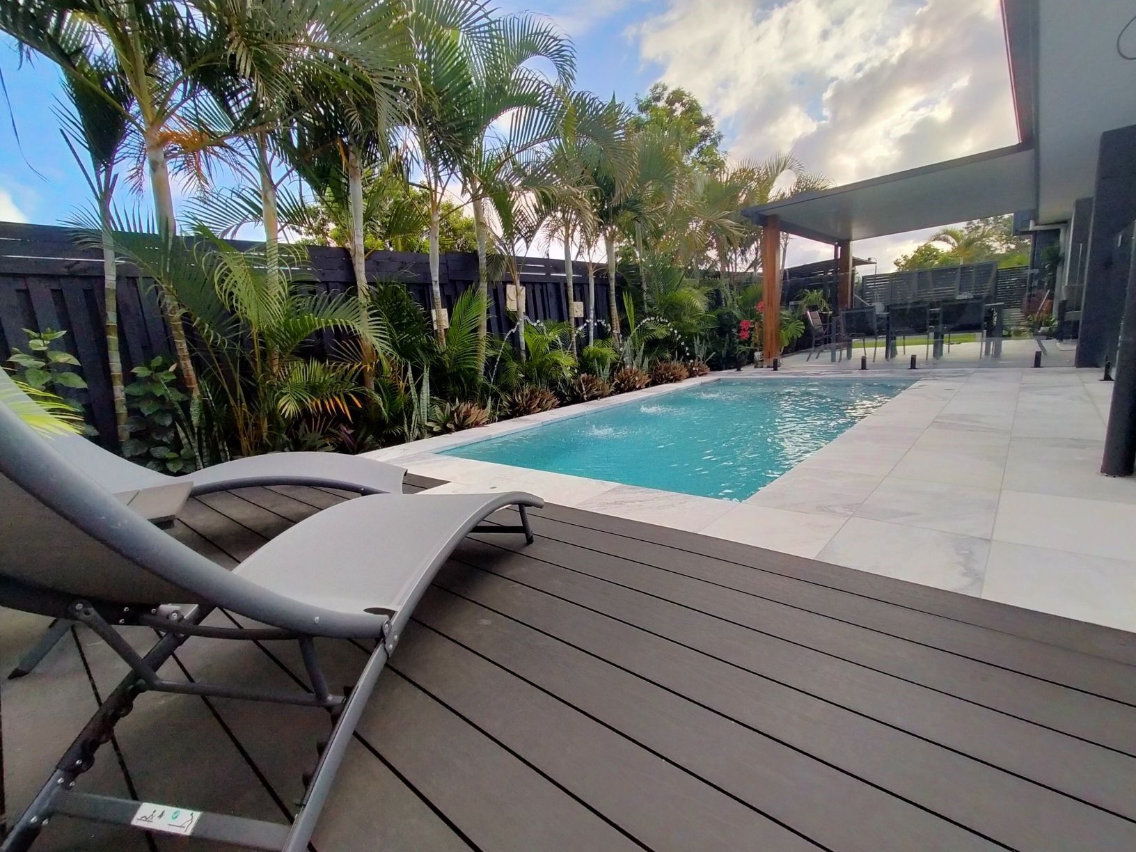 Pool with lounge chairs, surrounded by tropical plants, modern house in the background.