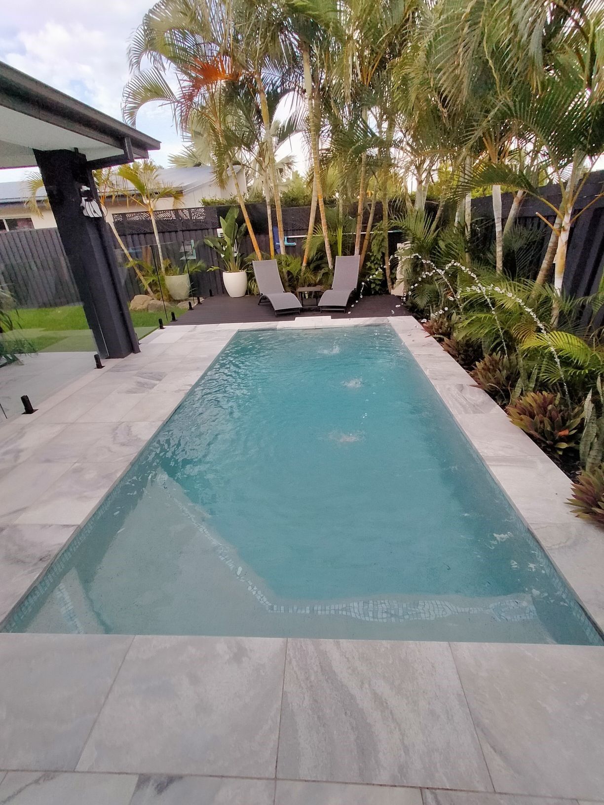 Rectangular pool surrounded by stone tiles. Palm trees, lounge chairs, and greenery in the background.