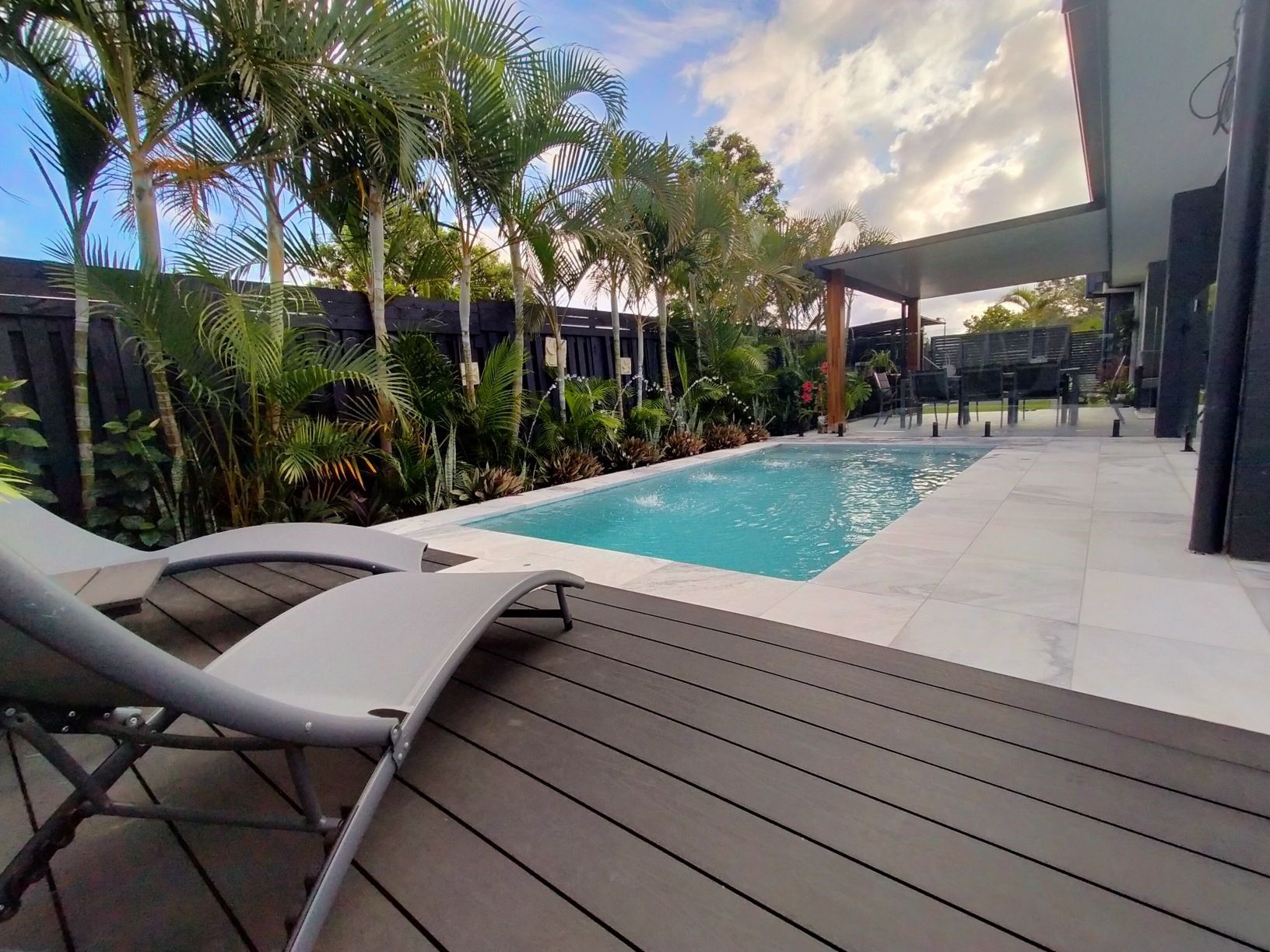 Poolside scene with lounge chairs, pool, lush greenery, and sunny sky.