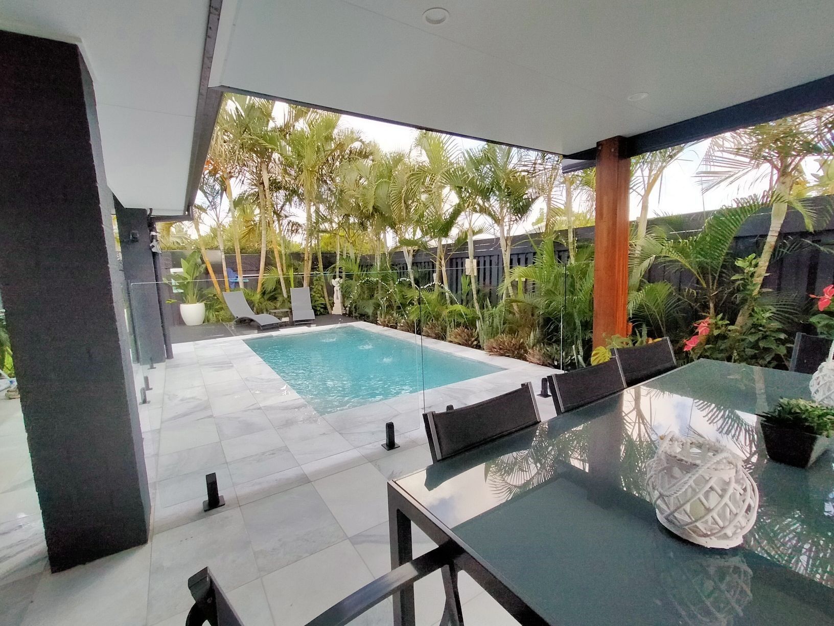 Patio with pool, dining table, and lush greenery. Gray and white tones, sunny outdoor setting.