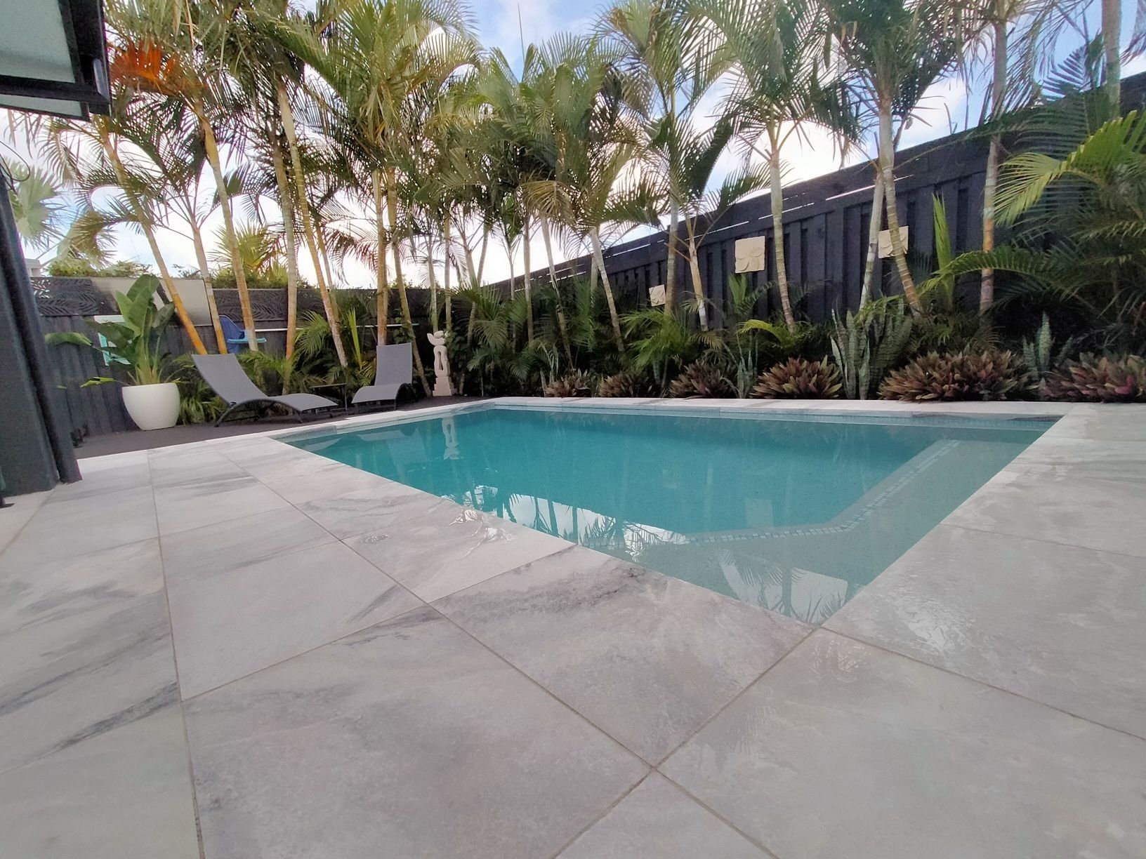 Pool area with gray tiling, clear water, palm trees, and lounge chairs.
