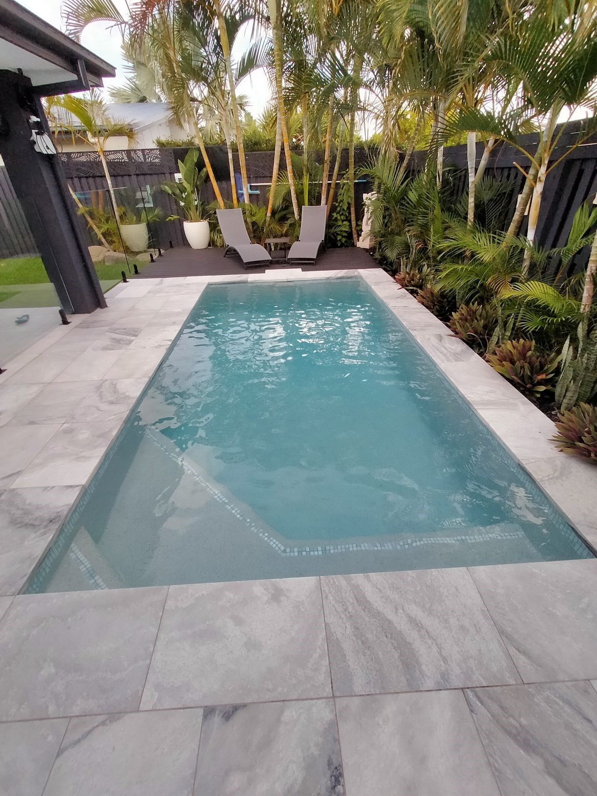 A rectangular pool with gray stone surround in a backyard setting with lounge chairs and landscaping.