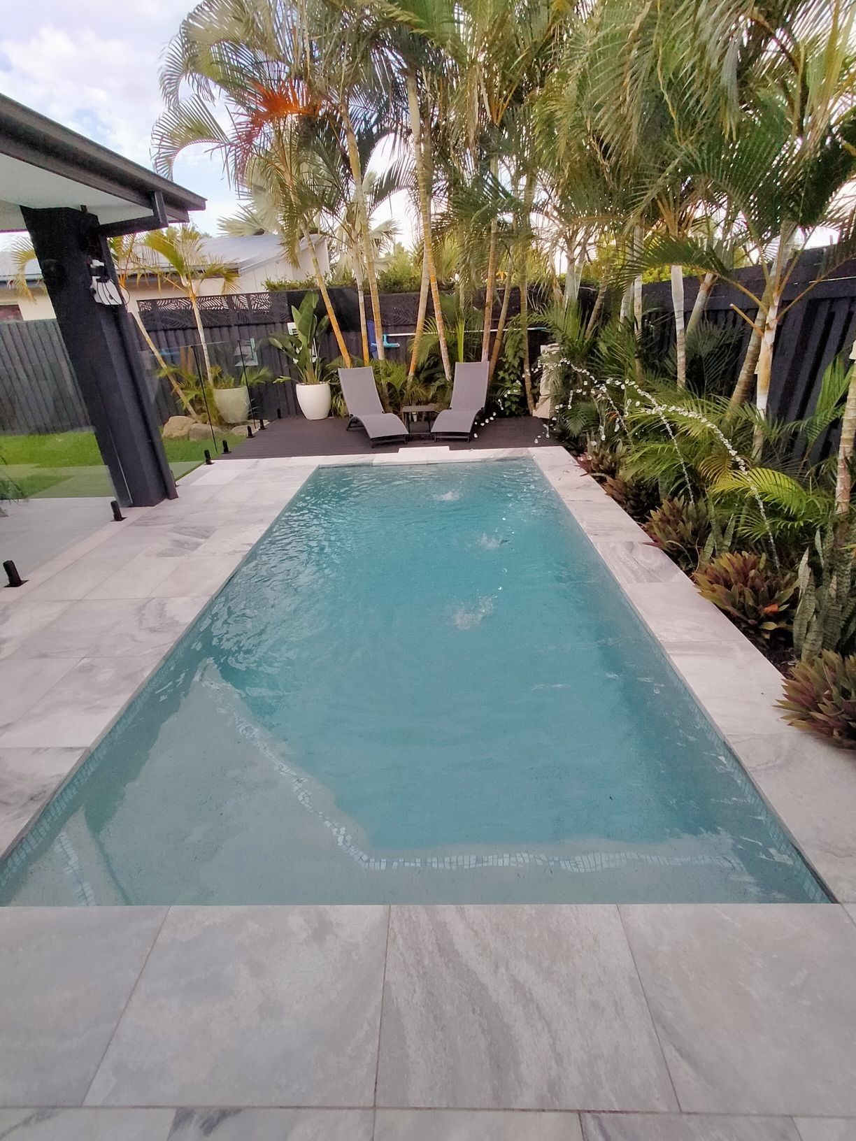 Rectangular pool with light blue water, surrounded by light gray tile, tropical foliage, and two chairs.