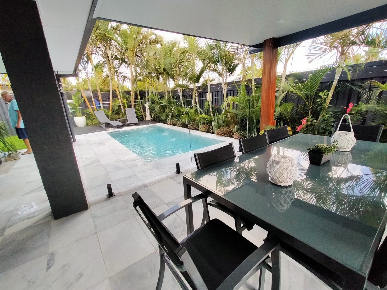 Poolside patio with dining table and pool, surrounded by lush greenery.