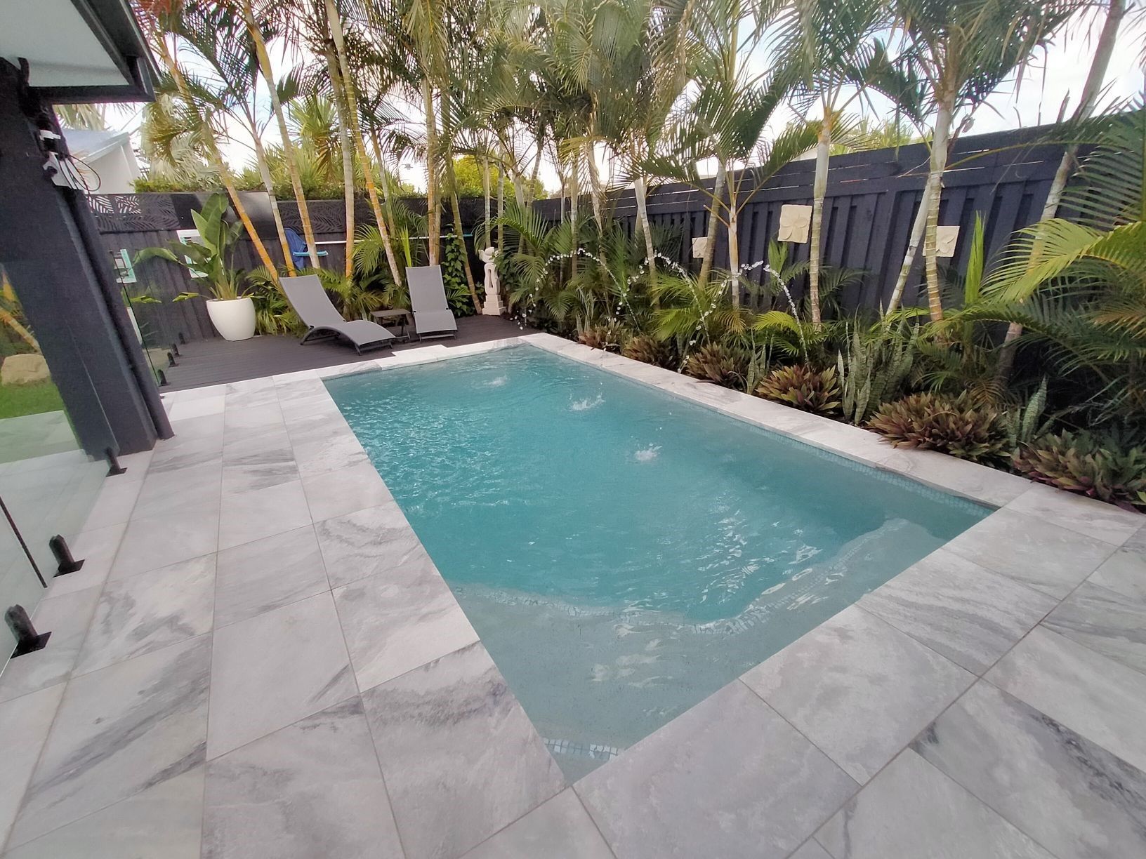 A rectangular pool in a backyard, surrounded by gray tiles, plants, and a black fence.