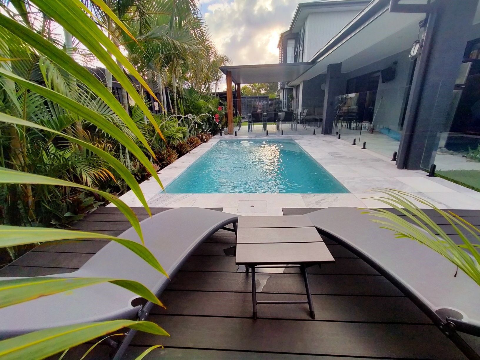 Lounge chairs on deck overlooking pool and modern home. Lush foliage, clear water, sunny day.