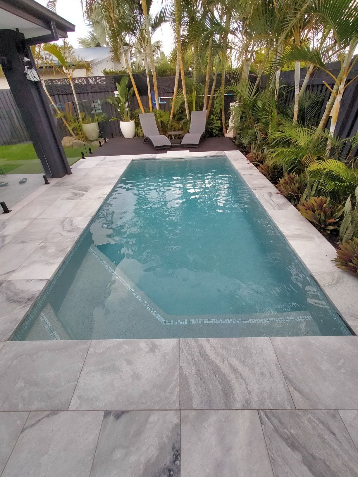 Rectangular pool with light blue water surrounded by gray stone tiles and lush greenery.
