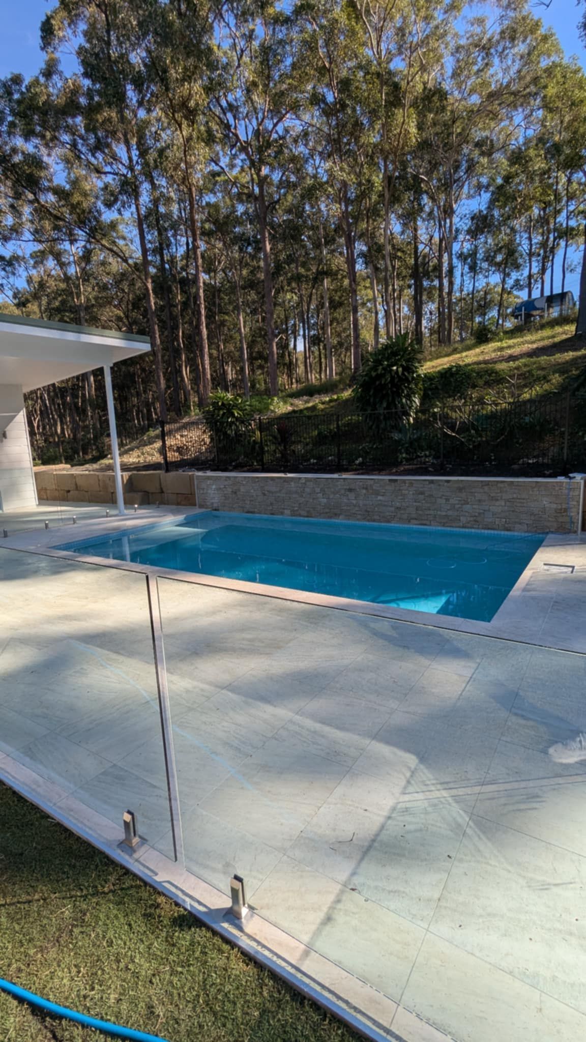 Swimming pool with clear glass fencing on a sunny day. Trees in the background.