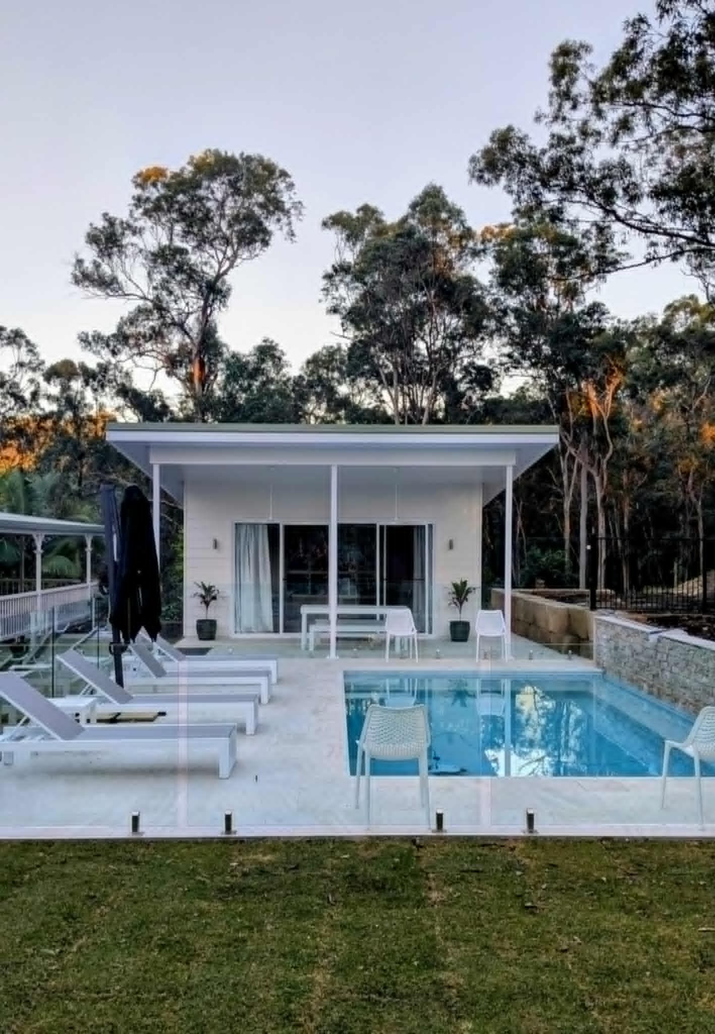 A modern white cabin with a pool, surrounded by trees and lounge chairs.