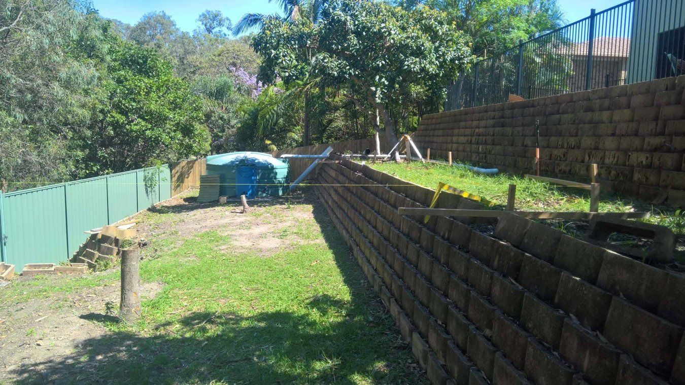 Green backyard with tiered retaining walls, green fence, trees, and blue tarp.