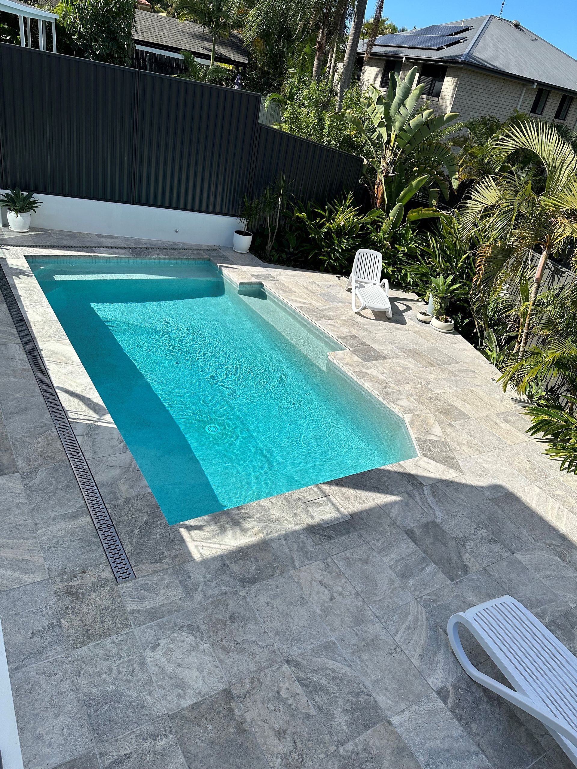 Rectangular swimming pool with turquoise water on a stone patio, bordered by plants and a fence.