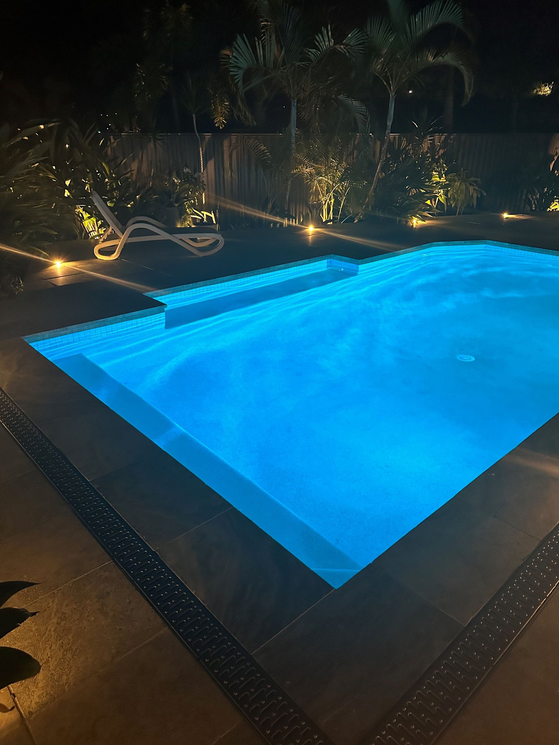 A blue-lit rectangular swimming pool at night with surrounding landscaping.