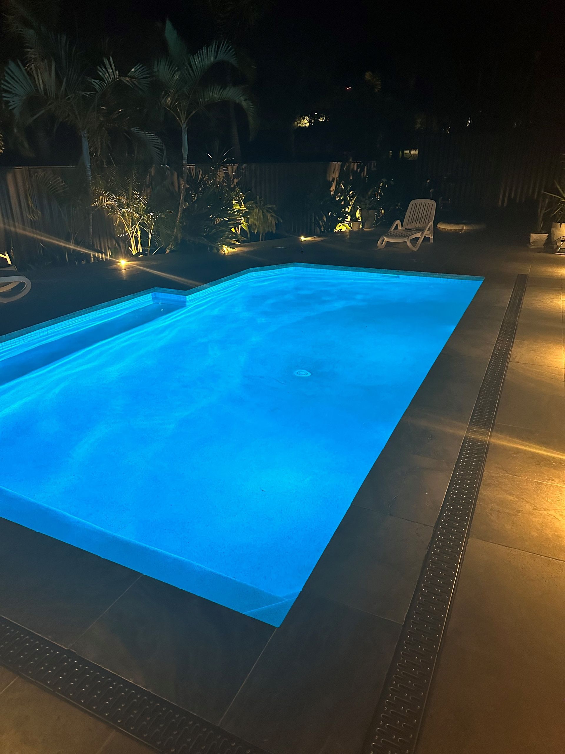 Nighttime illuminated pool with blue water, surrounded by a tiled patio and landscape lighting.